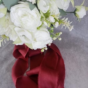 Könnte beinhalten: Ein Bouquet aus weißen Blumen, darunter Pfingstrosen und Rosen, mit grünem Laub und kleinen weißen Akzenten. Das Bouquet ist mit einem langen, fließenden weinroten Band gebunden, was einen romantischen Look erzeugt. Ideal für Hochzeiten.