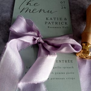 May include: A wedding menu card with the words "The menu" in elegant script. The card is sage green with the names Katie & Patrick and the date. A lavender ribbon is tied around the card.