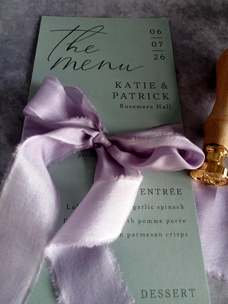 May include: A wedding menu card with the words "The menu" in elegant script. The card is sage green with the names Katie & Patrick and the date. A lavender ribbon is tied around the card.