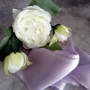 May include: A close-up of a white rose with green leaves and buds, resting on a folded lavender satin fabric. The fabric has a soft, textured appearance. The overall composition suggests a delicate and elegant theme.