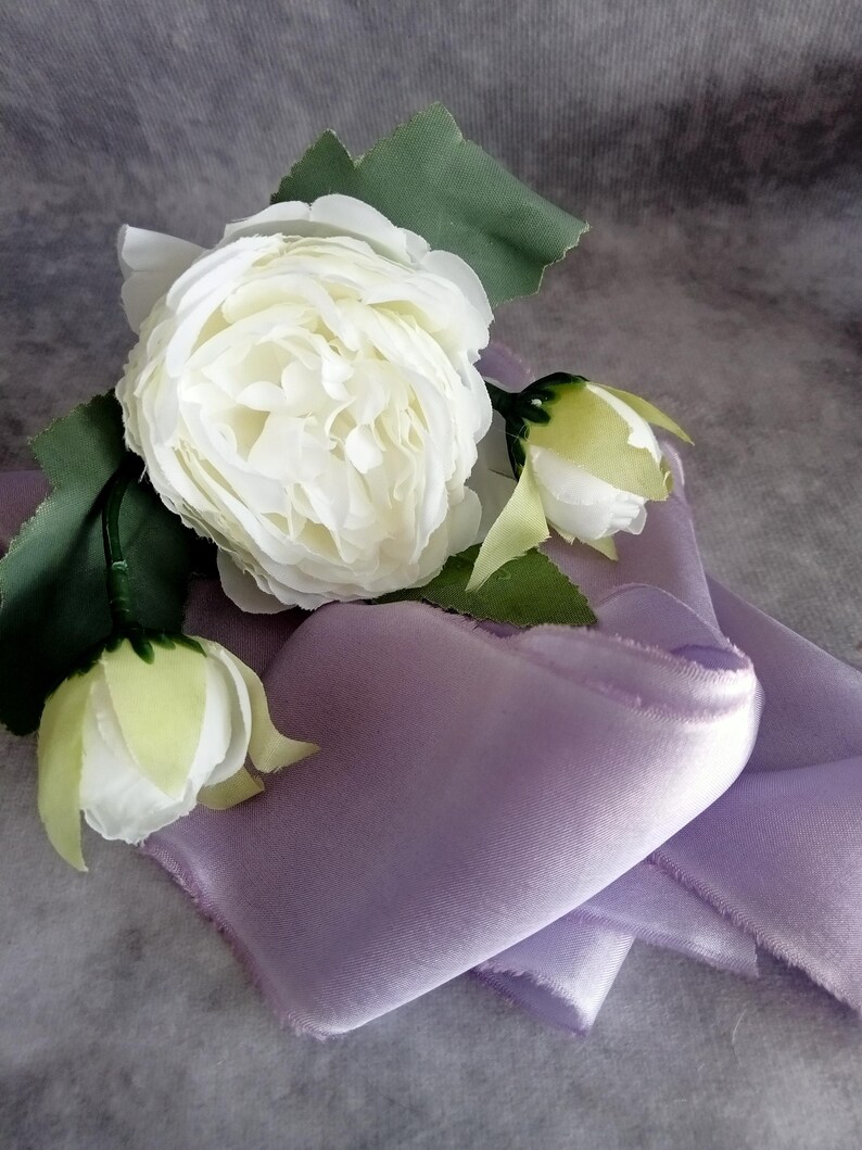 May include: A close-up of a white rose with green leaves and buds, resting on a folded lavender satin fabric. The fabric has a soft, textured appearance. The overall composition suggests a delicate and elegant theme.