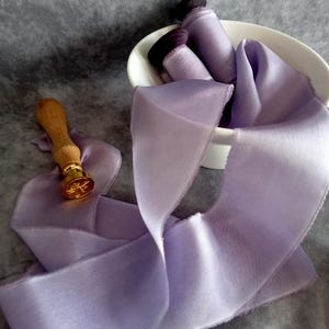 May include: A light purple silk ribbon with a frayed edge, flowing across a gray surface. A wooden handled wax seal with a gold emblem is next to the ribbon. Rolled ribbons are in a white bowl.