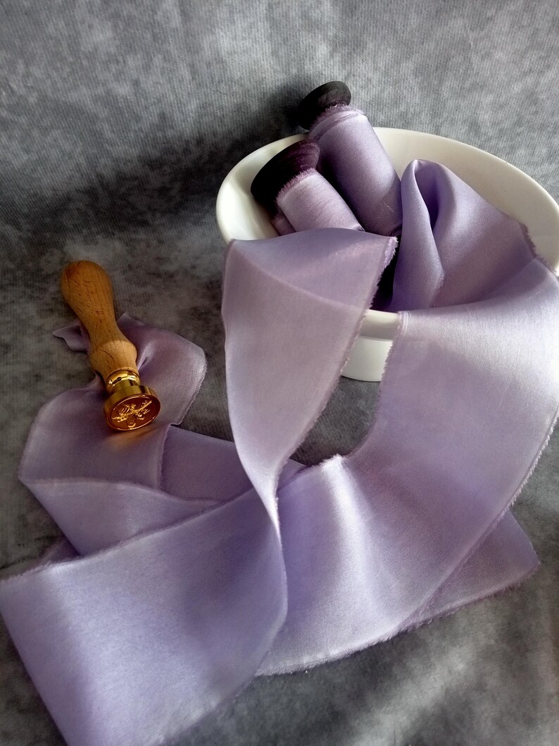 May include: A light purple silk ribbon with a frayed edge, flowing across a gray surface. A wooden handled wax seal with a gold emblem is next to the ribbon. Rolled ribbons are in a white bowl.
