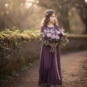 May include: A young person in a long, plum-colored dress and a floral crown holds a bouquet of lavender and purple flowers. The dress has long sleeves and a flowing skirt. The background is a blurred outdoor scene with a stone wall.
