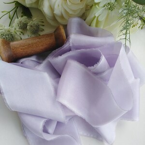 May include: A length of soft, lavender-colored silk ribbon with frayed edges, artfully arranged. A wooden spool rests nearby, with white roses and other flowers in the background. This ribbon is ideal for crafts and floral arrangements.