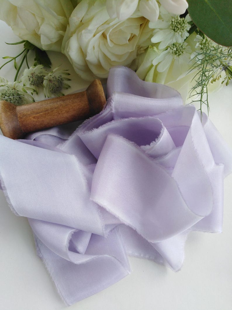 May include: A length of soft, lavender-colored silk ribbon with frayed edges, artfully arranged. A wooden spool rests nearby, with white roses and other flowers in the background. This ribbon is ideal for crafts and floral arrangements.