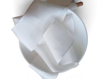 Op de afbeelding: Een lang, gebroken wit stoffen lint met rafelige randen is opgerold en gedrapeerd in een eenvoudige witte keramische kom. Het lint heeft een gladde, matte textuur en is gedeeltelijk gewikkeld rond een kleine, lichtgekleurde houten klos.