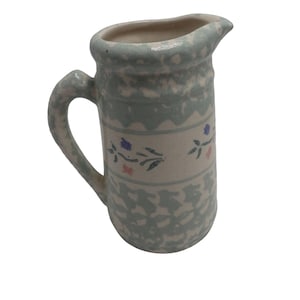 May include: A small, ceramic pitcher with a handle and spout. The pitcher is primarily off-white with a light blue floral pattern and accents. The handle and rim are also light blue. The pitcher has a classic, rustic design.