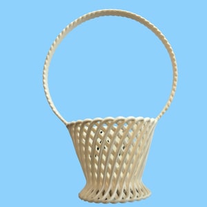 May include: Off-white woven basket with a curved handle. The basket has a flared base and a lattice design. The handle is a thick, rounded shape. The basket is set against a light blue background.
