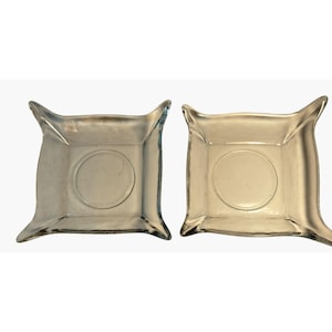 May include: Two clear glass square dishes with rounded corners and a circular indentation in the center. The dishes have a slightly raised rim and are set against a white background.