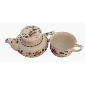 May include: A white ceramic tea set comprising a teapot with a lid and a matching teacup. Both items are decorated with a delicate floral pattern featuring pink roses and blue flowers, complemented by thin green leaves and a fine gold rim on the lid.