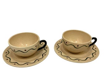 May include: Two beige ceramic teacups with matching saucers. Each cup has a black handle and is decorated with a black wavy line and red dots. The saucers have a matching design. A charming set for serving tea.