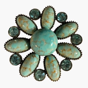 May include: A circular brooch featuring a large central round turquoise-colored stone with brown veining, surrounded by six oval turquoise-colored stones with brown veining and six smaller round light blue faceted crystals in a flower-like pattern.