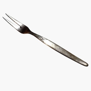 Puede incluir: Un tenedor de plata vintage con dos púas. El mango tiene un diseño floral ornamentado cerca de la cabeza del tenedor. El tenedor está en ángulo diagonalmente contra un fondo blanco, mostrando su apariencia envejecida y deslustrada.
