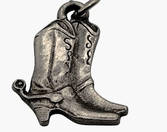 Puede incluir: Un dije de metal plateado con forma de un par de botas vaqueras. Las botas presentan detalles de costuras decorativas en relieve en las cañas y una pequeña espuela unida al talón de la bota trasera. Una argolla circular está sujeta en la parte superior.