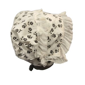 May include: A white bonnet with a floral pattern in dark brown. The bonnet features a ruffled brim and a tie closure. The fabric has a soft, slightly textured appearance, and the floral design is evenly distributed across the surface.
