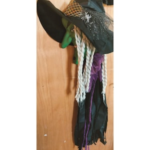 May include: A Halloween witch decoration with a green face, long white braids, and a black hat. The hat is decorated with a spider and netting. Purple ribbon and black fabric hang from the witch's neck.