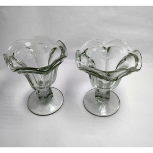 Puede incluir: Dos copas de helado de cristal transparente. Cada copa tiene un borde festoneado, un tallo facetado y una base redonda. El cristal transparente permite el paso de la luz. Estas copas de estilo vintage son adecuadas para servir postres.