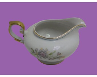 May include: A white porcelain creamer with a gold rim and floral design. The creamer has a rounded body and a curved handle.