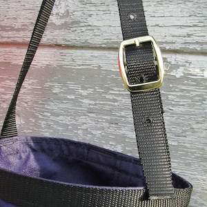 Puede incluir: Primer plano de un bolso tote morado y negro con una correa negra y una hebilla dorada. El interior del bolso es de color morado oscuro, y el exterior presenta una banda negra. La correa est&aacute; hecha de un material tejido duradero.