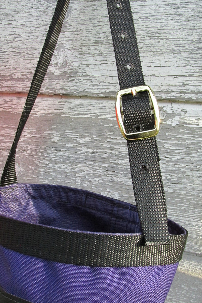 Puede incluir: Primer plano de un bolso tote morado y negro con una correa negra y una hebilla dorada. El interior del bolso es de color morado oscuro, y el exterior presenta una banda negra. La correa est&aacute; hecha de un material tejido duradero.