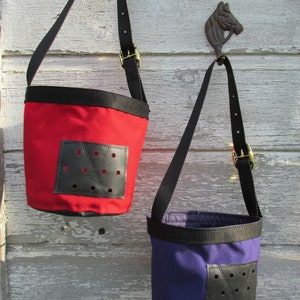 Puede incluir: Dos peque&ntilde;as bolsas en forma de cubo, rojas y moradas, colgadas de ganchos. Cada bolsa tiene una correa negra, un parche de cuero negro con agujeros y una hebilla dorada. Las bolsas cuelgan de una superficie de madera blanca desgastada.