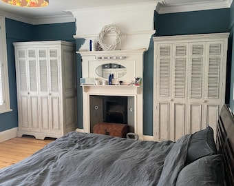 Può includere: Una camera da letto con un copriletto grigio e persiane bianche sulle porte dell'armadio. Le pareti sono dipinte di un colore blu turchese. C'è un camino con un camino in legno e una ghirlanda bianca appesa sopra.