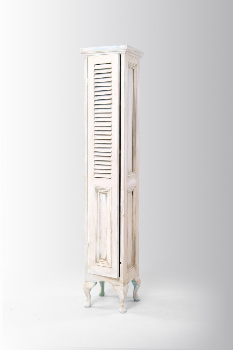 Pu&ograve; includere: Un armadio in legno bianco con una porta a battente e gambe decorative. L'armadio ha una finitura di vernice invecchiata ed &egrave; un design alto e stretto.