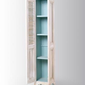 Pu&ograve; includere: Un armadio in legno bianco con un interno blu chiaro e quattro gambe. La porta dell'armadio &egrave; aperta, rivelando tre ripiani.