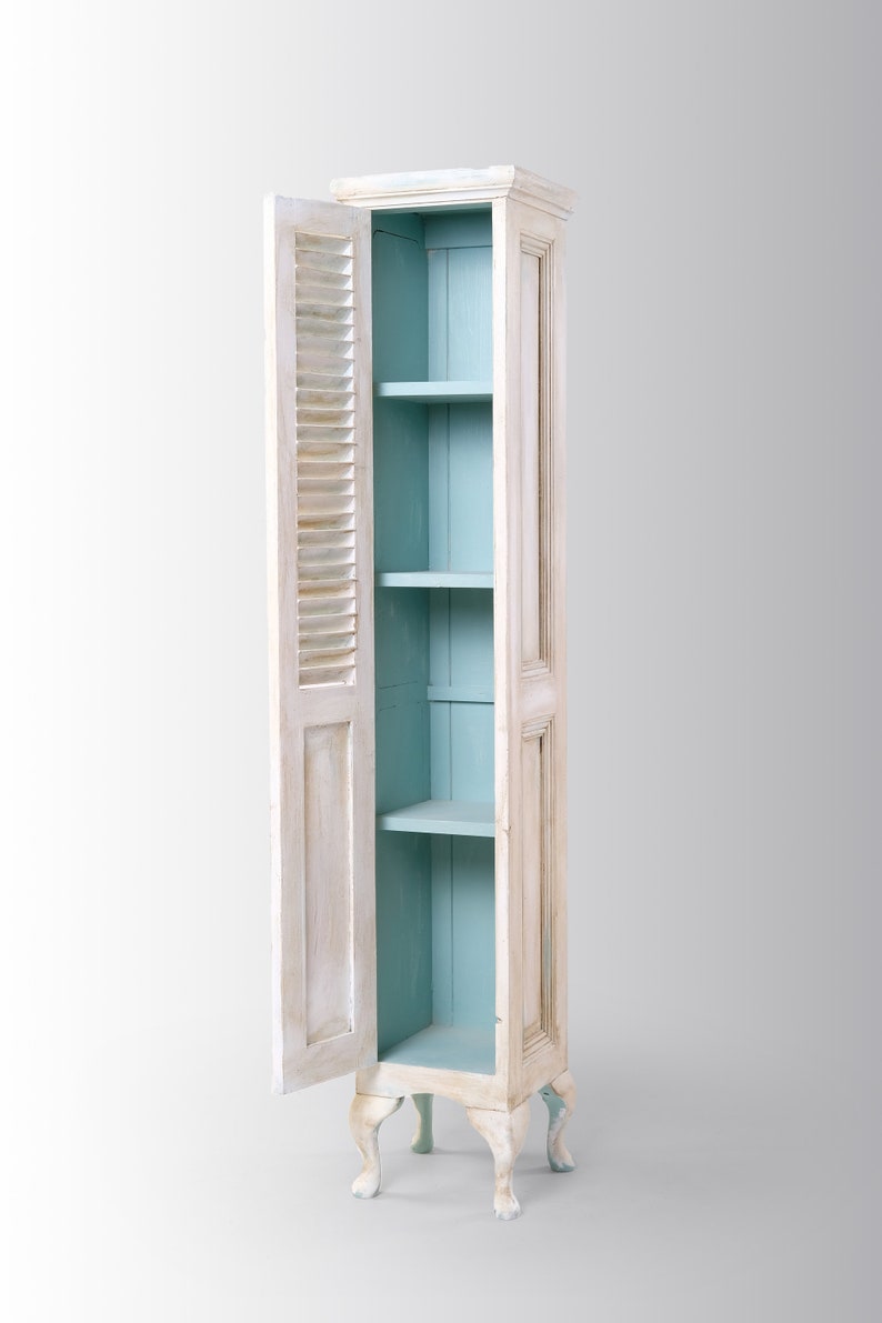 Pu&ograve; includere: Un armadio in legno bianco con un interno blu chiaro e quattro gambe. La porta dell'armadio &egrave; aperta, rivelando tre ripiani.