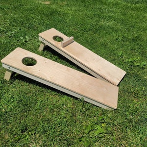 May include: Two wooden cornhole boards with a natural wood finish and white trim. The boards are set up on a green grassy surface.