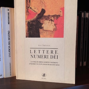 Może przedstawiać: Książka zatytułowana "LETTERE, NUMERI, DEI" autorstwa Guya Trévoux. Okładka przedstawia ilustrację dwóch postaci w czerwonych i czarnych szatach. Książka jest w języku włoskim i znajduje się na półce z innymi książkami.