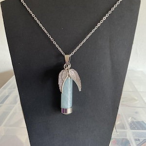K&ouml;nnte beinhalten: Halskette mit silberfarbenem Anh&auml;nger. Der Anh&auml;nger hat einen hellblauen Kristall in einem silbernen Metallrohr und silberne Engelsfl&uuml;gel. Die Halskette wird auf einem schwarzen St&auml;nder pr&auml;sentiert.