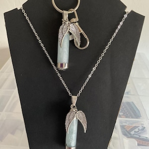 K&ouml;nnte beinhalten: Ein Halsketten- und Schl&uuml;sselanh&auml;nger-Set mit einem hellblauen zylindrischen Anh&auml;nger mit silberfarbenen Fl&uuml;gelakzenten. Die Halskette hat eine zarte Kette, und der Schl&uuml;sselanh&auml;nger enth&auml;lt einen Karabiner und einen Ring. Die Artikel werden auf einem schwarzen St&auml;nder pr&auml;sentiert.