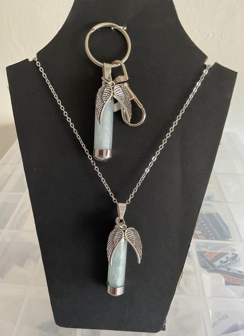 K&ouml;nnte beinhalten: Ein Halsketten- und Schl&uuml;sselanh&auml;nger-Set mit einem hellblauen zylindrischen Anh&auml;nger mit silberfarbenen Fl&uuml;gelakzenten. Die Halskette hat eine zarte Kette, und der Schl&uuml;sselanh&auml;nger enth&auml;lt einen Karabiner und einen Ring. Die Artikel werden auf einem schwarzen St&auml;nder pr&auml;sentiert.