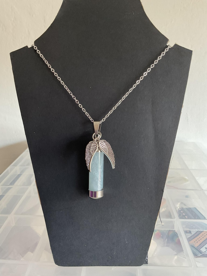 K&ouml;nnte beinhalten: Halskette mit silberfarbenem Anh&auml;nger. Der Anh&auml;nger hat einen hellblauen Kristall in einem silbernen Metallrohr und silberne Engelsfl&uuml;gel. Die Halskette wird auf einem schwarzen St&auml;nder pr&auml;sentiert.