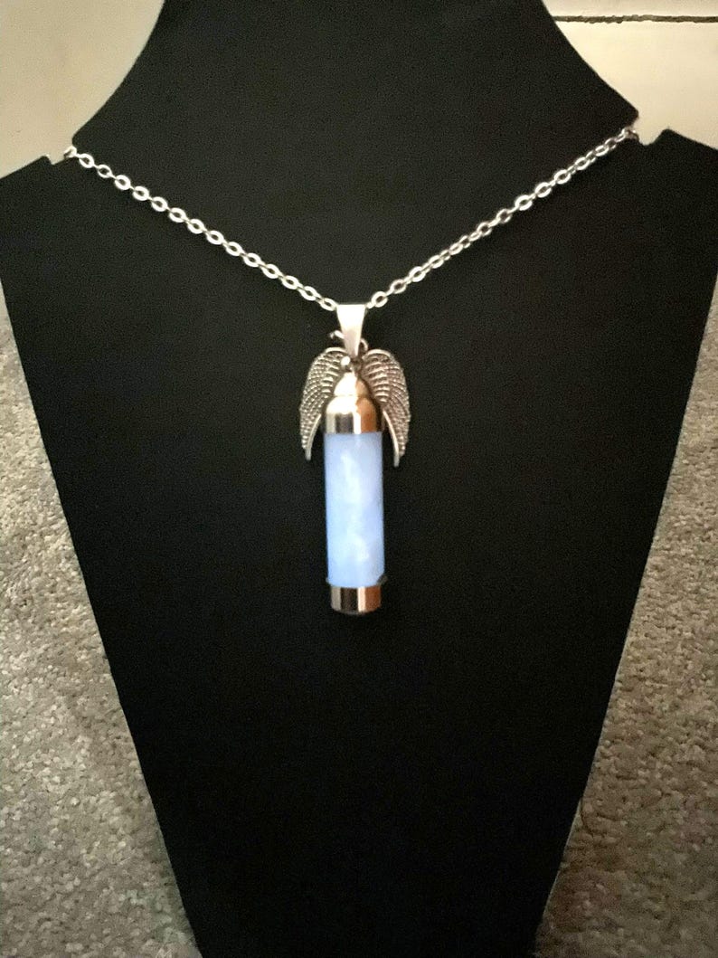 K&ouml;nnte beinhalten: Halskette mit silberfarbenem Anh&auml;nger. Der Anh&auml;nger hat einen hellblauen zylindrischen Stein und silberfarbene Fl&uuml;gel. Pr&auml;sentiert auf einem schwarzen St&auml;nder.
