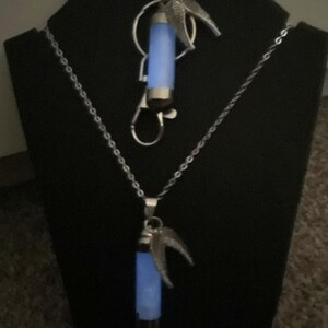 K&ouml;nnte beinhalten: Zwei silberfarbene Halsketten mit leuchtend blauen Anh&auml;ngern. Jeder Anh&auml;nger ist zylindrisch mit silbernen Akzenten und einem dekorativen Fl&uuml;geldesign. Eine Halskette ist ein Anh&auml;nger, die andere eine Halskette.