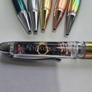 May include: A selection of pens with metallic barrels in various colours, including green, gold, and iridescent. One pen has a clear barrel with a "Supernatural" design and a silver clip. The pens are arranged on a white surface.
