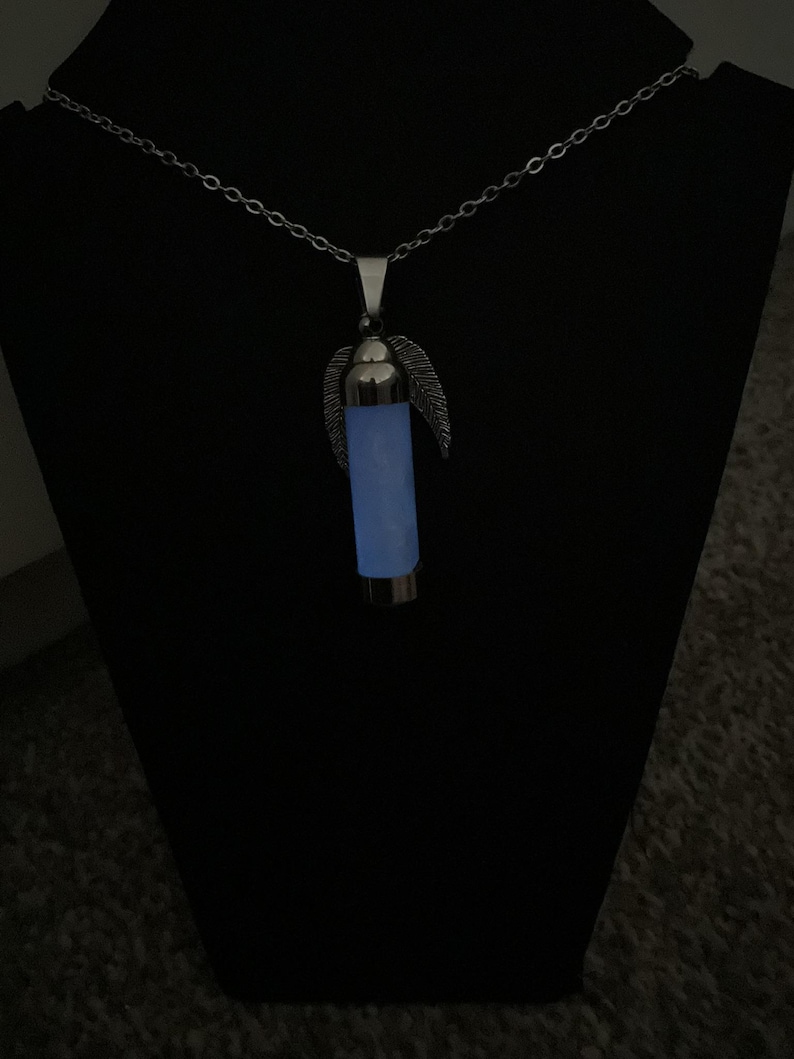 K&ouml;nnte beinhalten: Halskette mit silberfarbenem Anh&auml;nger. Der Anh&auml;nger hat ein leuchtendes blaues zylindrisches Design mit federartigen Details. Der Anh&auml;nger h&auml;ngt an einer filigranen Kette vor dunklem Hintergrund.