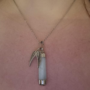K&ouml;nnte beinhalten: Halskette mit silberfarbenem Anh&auml;nger. Der Anh&auml;nger hat einen hellblauen, zylindrischen Stein und dekorative silberne Fl&uuml;gel. Die Halskette h&auml;ngt an einer filigranen Kette. Der Hintergrund ist rosa.