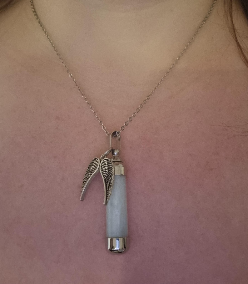 K&ouml;nnte beinhalten: Halskette mit silberfarbenem Anh&auml;nger. Der Anh&auml;nger hat einen hellblauen, zylindrischen Stein und dekorative silberne Fl&uuml;gel. Die Halskette h&auml;ngt an einer filigranen Kette. Der Hintergrund ist rosa.