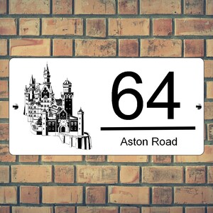 May include: White rectangular house number sign with the number "64" in black, and the words "Aston Road" below. A black and white illustration of a castle is on the left side. The sign is mounted on a brick wall.