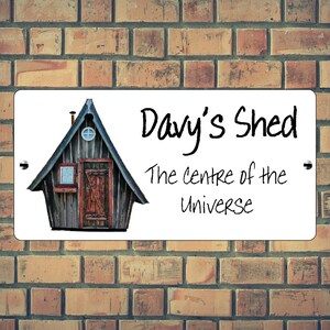 May include: White sign with a cartoon drawing of a wooden shed with a red door and a small window. The text on the sign reads "Davy's Shed The centre of the Universe".
