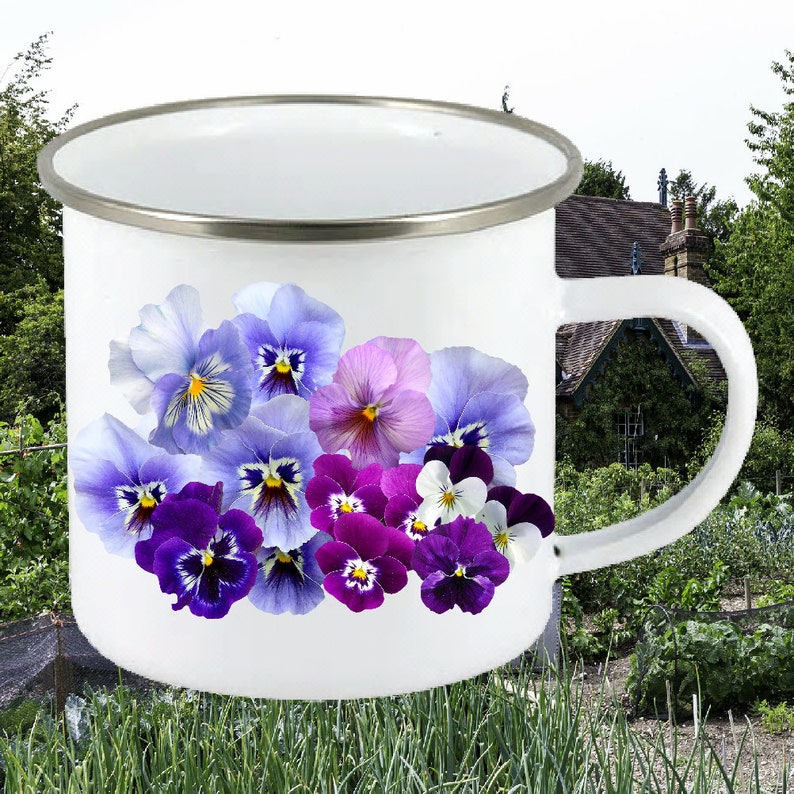 Puede incluir: Taza de esmalte blanco con un dise&ntilde;o floral colorido con pensamientos morados, rosas y azules. La taza tiene un borde plateado y un asa.