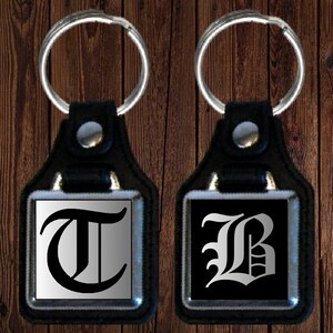 May include: Two black leather keychains with silver metal frames. The keychain on the left has a silver background with a black letter "T" in a medieval font. The keychain on the right has a black background with a silver letter "B" in a medieval font.