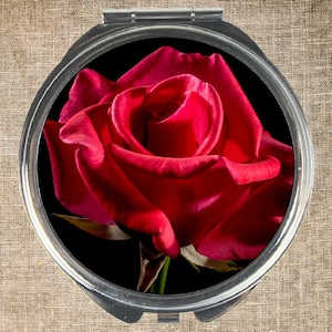 May include: A compact mirror with a single red rose on a black background. The rose is in full bloom and has a deep red color.