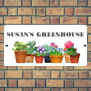 May include: White sign with black text that reads "SUSAN'S GREENHOUSE". The sign features a row of potted plants with various flowers and foliage, including a cactus, ivy, and a geranium.