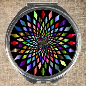 May include: A round, silver compact mirror with a colourful, geometric design. The design features a spiral of diamonds in various shades of red, orange, yellow, green, blue, purple, and pink on a black background.