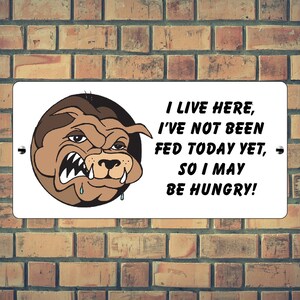 May include: A rectangular white metal sign with a cartoon bulldog illustration and the text "I LIVE HERE, I'VE NOT BEEN FED TODAY YET, SO I MAY BE HUNGRY!" The sign is mounted on a brick wall.